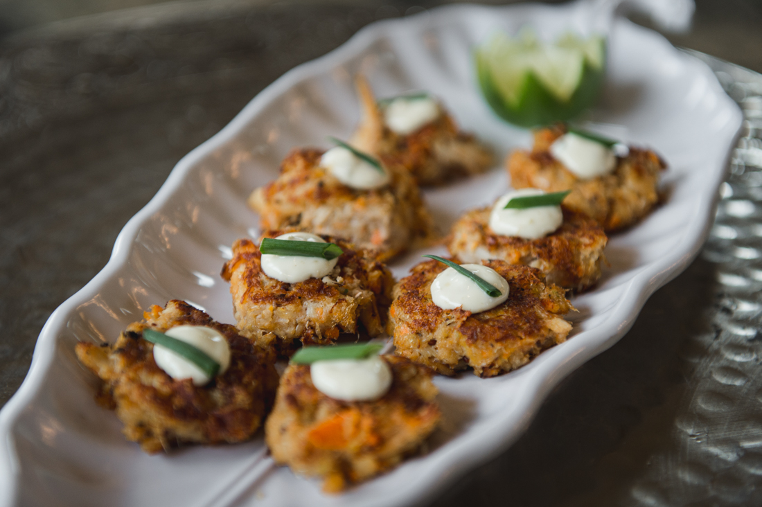 Mini crab cakes topped with a dollop of white sauce and a sliver of green onion, served on a white platter.