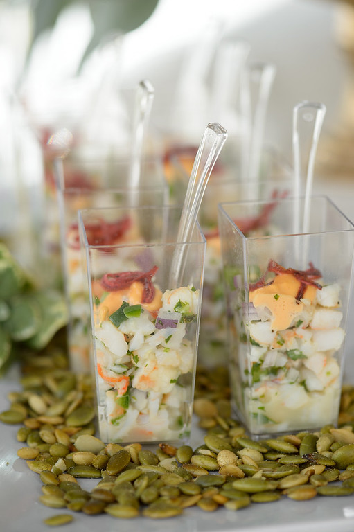 Seafood ceviche shooters in mini square plastic cups topped with a dollop of creamy orange sauce and thin red garnishes, arranged on a bed of pepitas (pumpkin seeds).