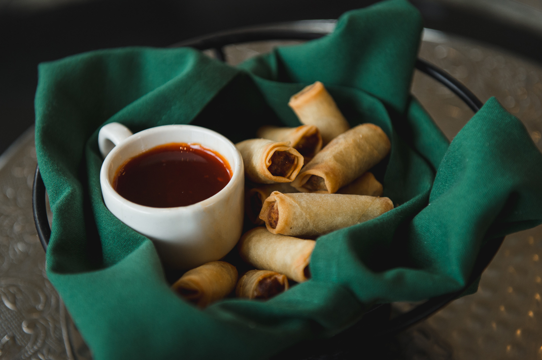 Crispy spring rolls served in a basket lined with a green napkin, accompanied by a small white cup of sweet and sour dipping sauce.