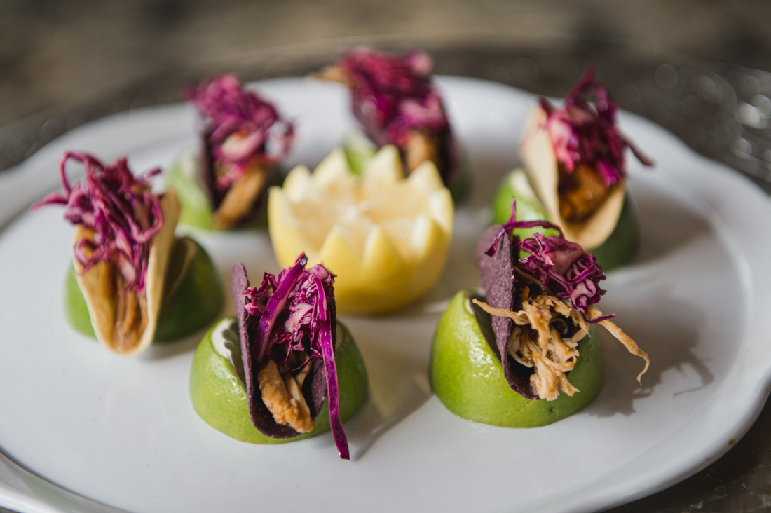 Mini tacos filled with shredded meat and red cabbage slaw, resting on lime wedges on a white platter around a carved lemon garnish.