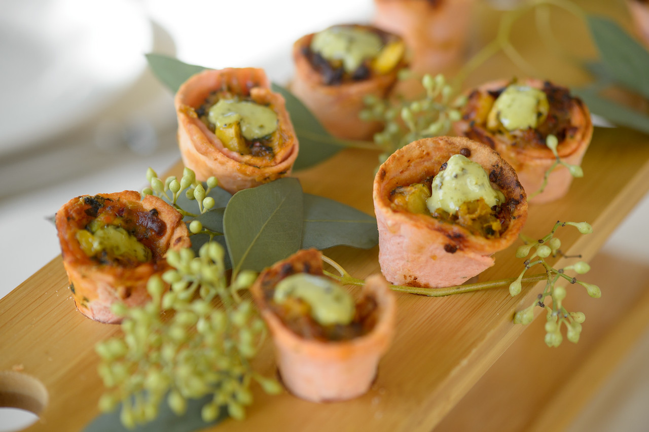 Mini tortilla cups filled with a savory mixture and topped with a dollop of green sauce, arranged on a wooden board with green leaves and berries.