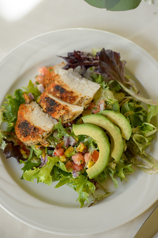 Grilled chicken salad with mixed greens, tomato corn salsa, and fresh avocado slices, served on a white plate.