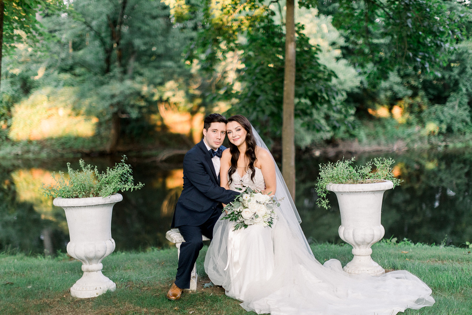 Bride and groom posing on a bench between two planters by a scenic riverbank.