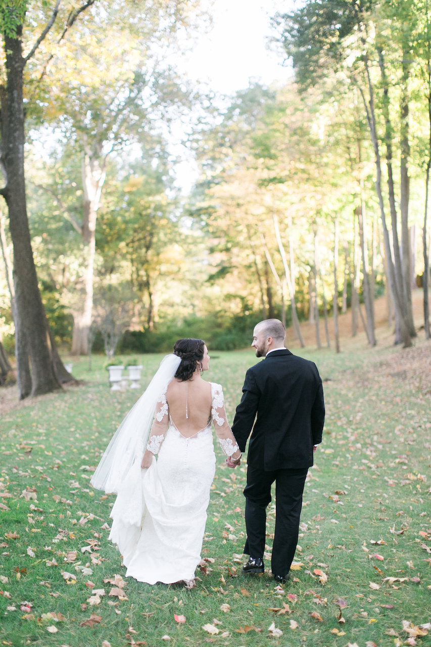 Bride and groom hold hands and walk away on a grassy lawn covered with scattered leaves, surrounded by trees.