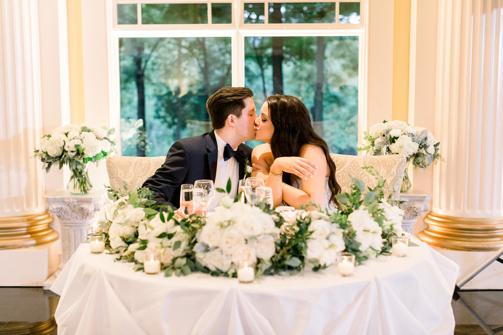 Bride and groom kissing while seated at a sweetheart table decorated with white floral garlands and candles.