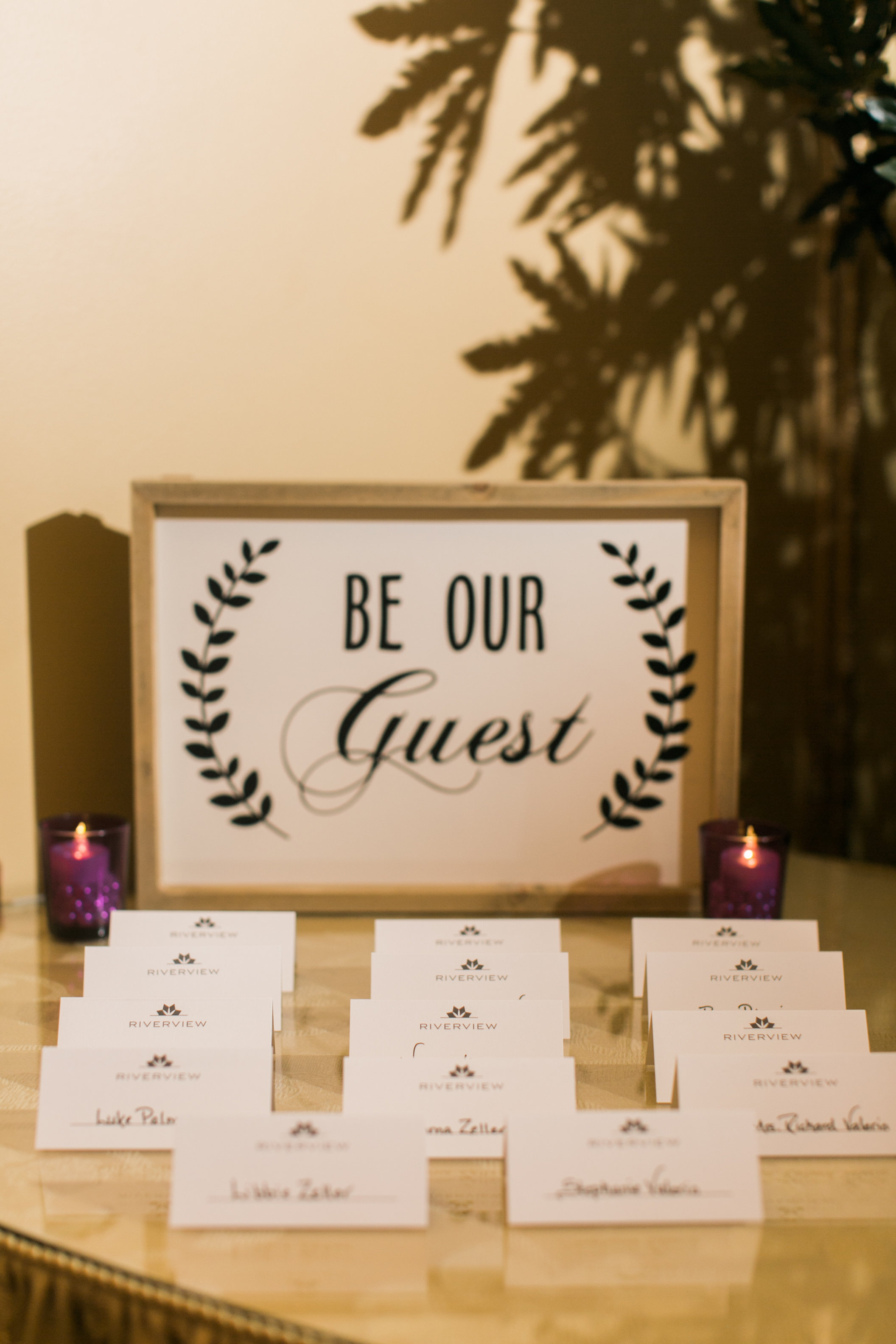 Wedding reception welcome table displaying a framed "Be Our Guest" sign, flanked by purple votive candles, with rows of white place cards arranged in front.