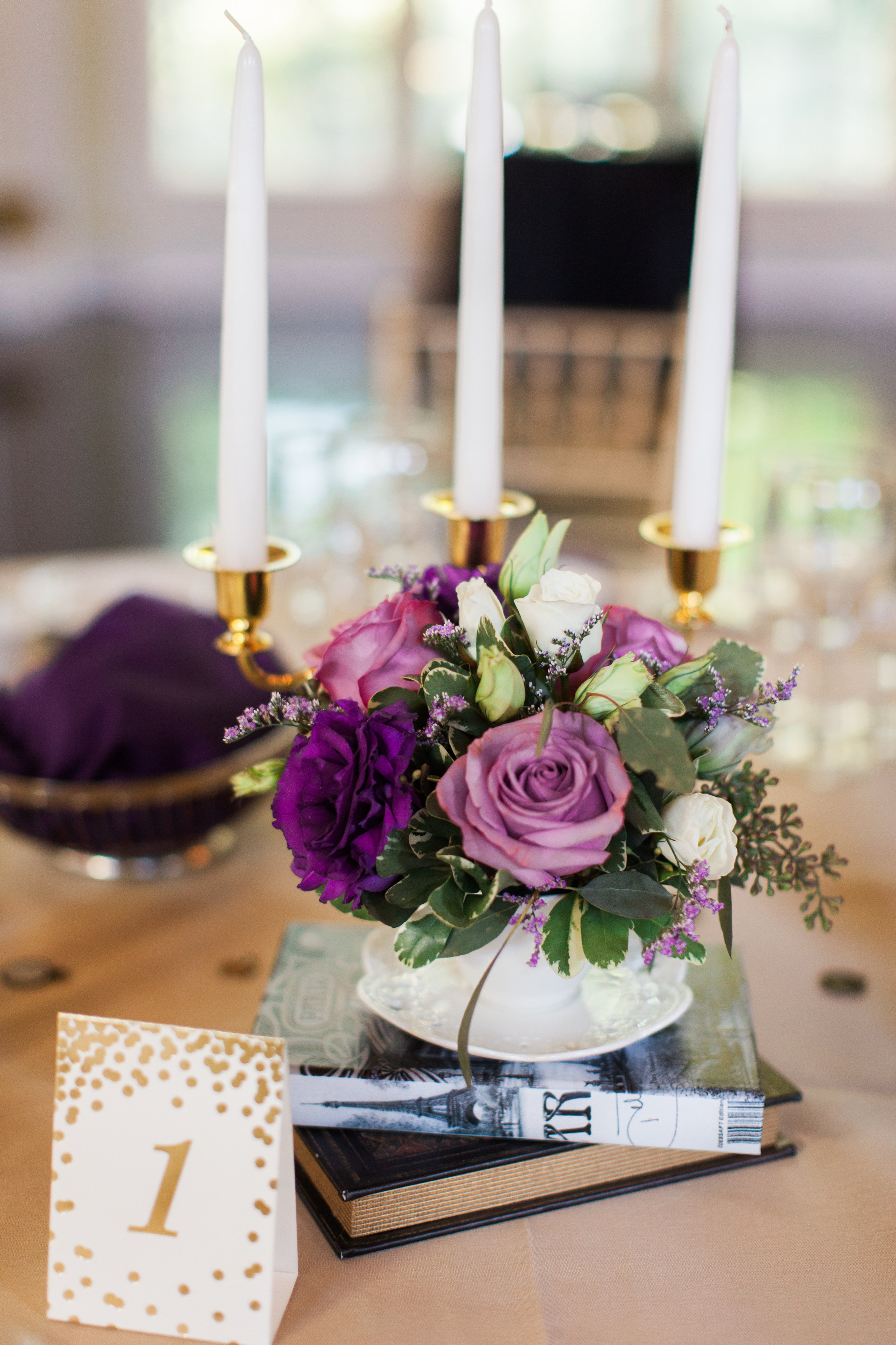 Wedding reception table centerpiece featuring purple and white roses in a teacup resting on a stack of books, next to a gold-candlestick candelabra and a number "1" table card.