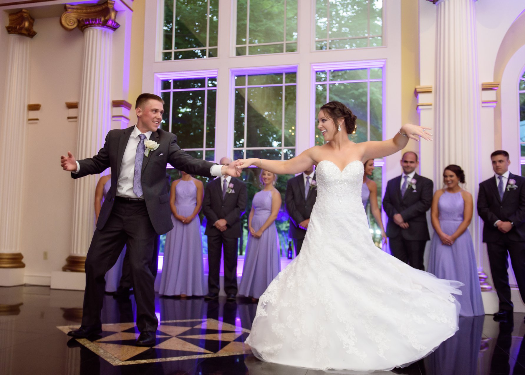 Groom spins his bride during their first dance on a reflective ballroom floor, surrounded by their bridal party in purple dresses and suits.