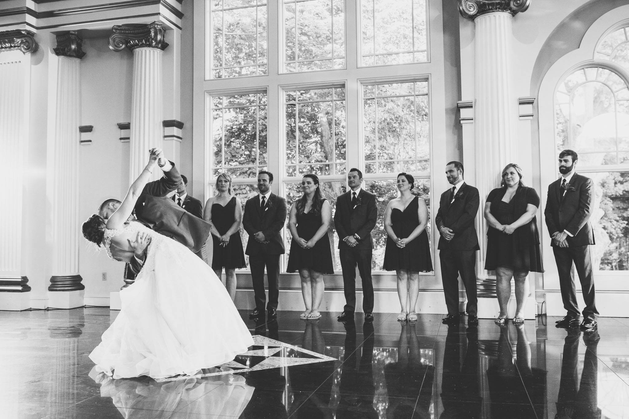 Black and white photo of a groom dipping his bride during their first dance on a reflective ballroom floor as wedding guests watch.