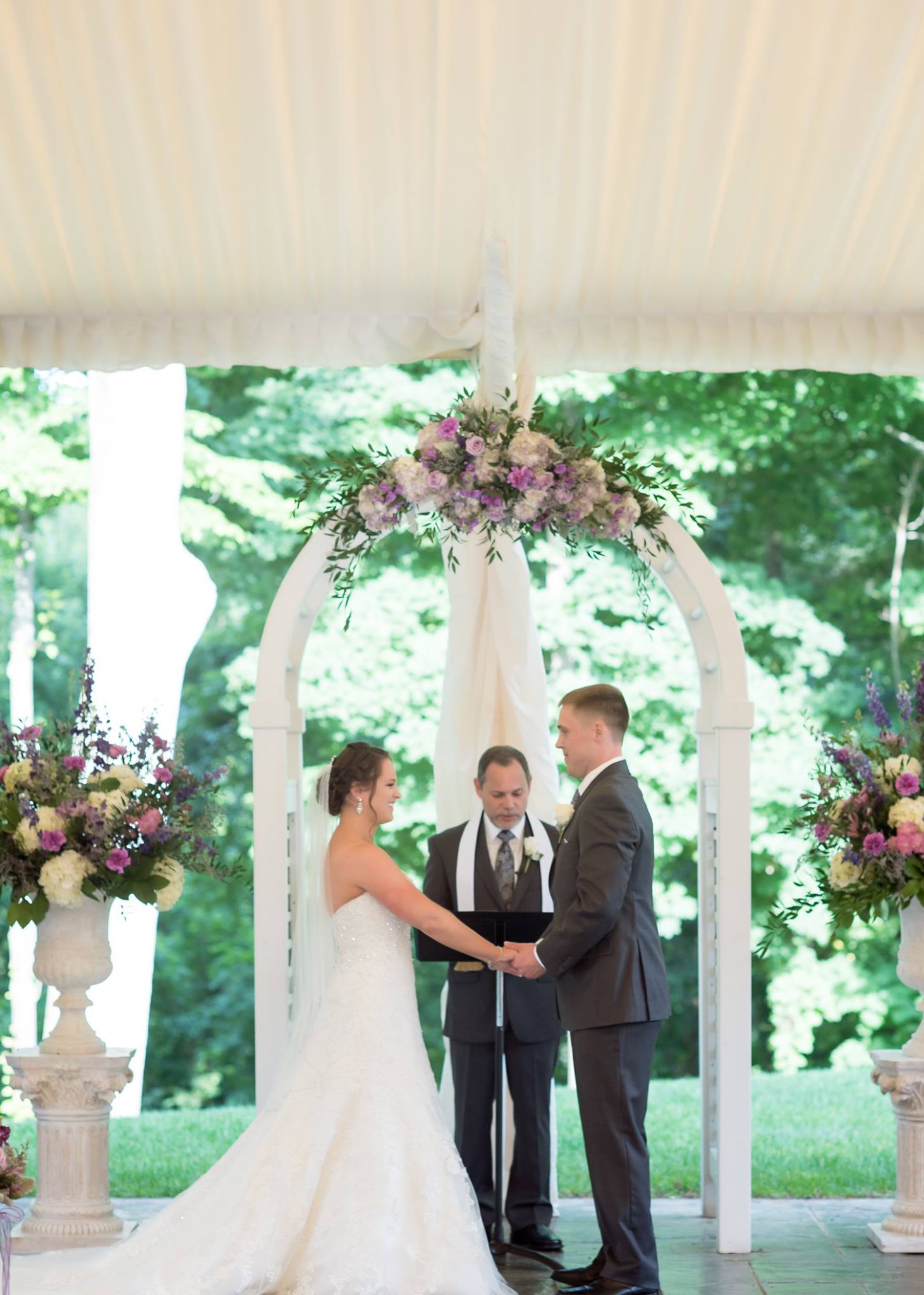 Bride and groom holding hands at the altar during an outdoor wedding ceremony under a floral arch.