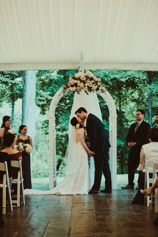 Bride and groom kissing under a white floral arch during an outdoor wedding ceremony.