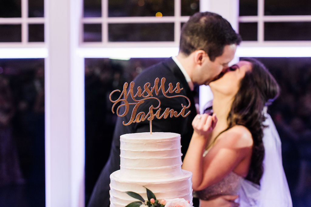 Bride and groom kissing behind a white wedding cake topped with a personalized script "Mr. & Mrs. Iasimi" cake topper.