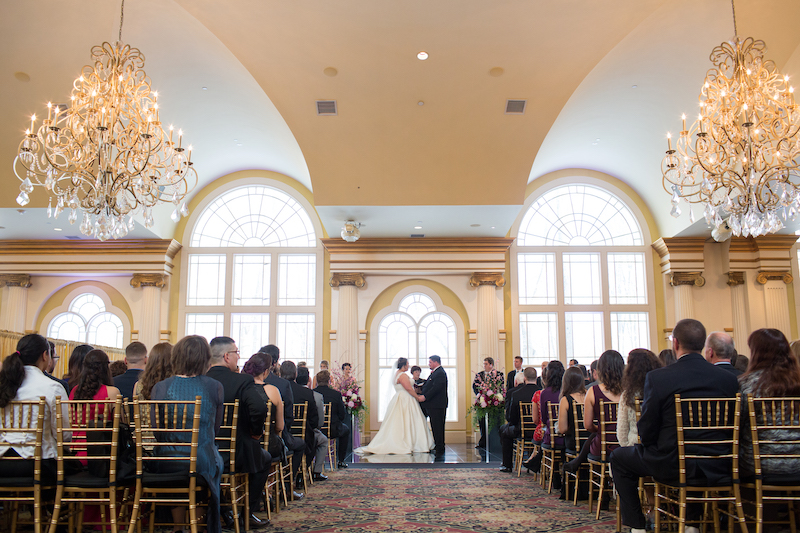 Bride and groom hold hands at the altar during an indoor ceremony in a grand ballroom with large windows.