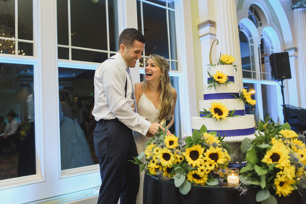 Bride and groom laughing together while cutting a multi-tier wedding cake decorated with sunflowers.
