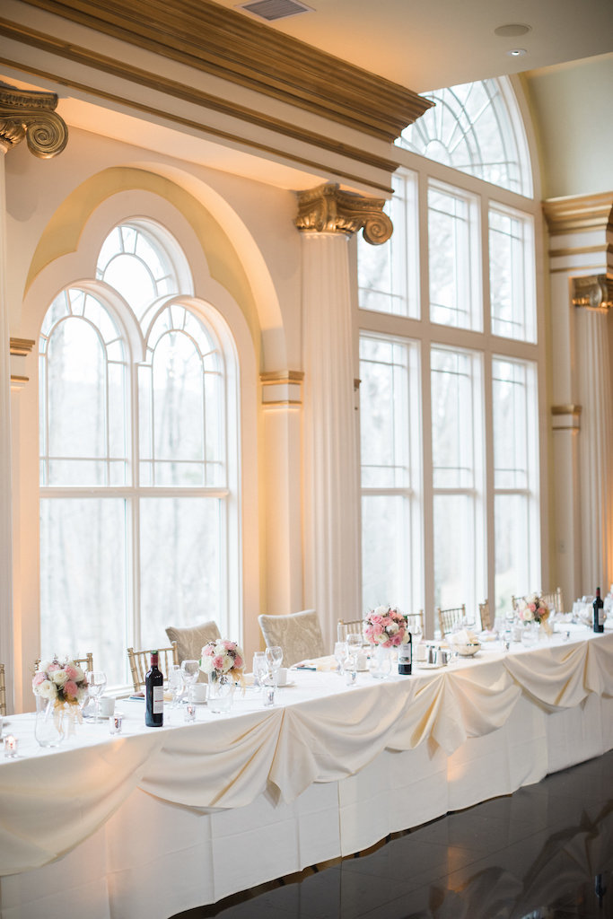 Long wedding head table decorated with pink floral arrangements and white drapes next to windows.