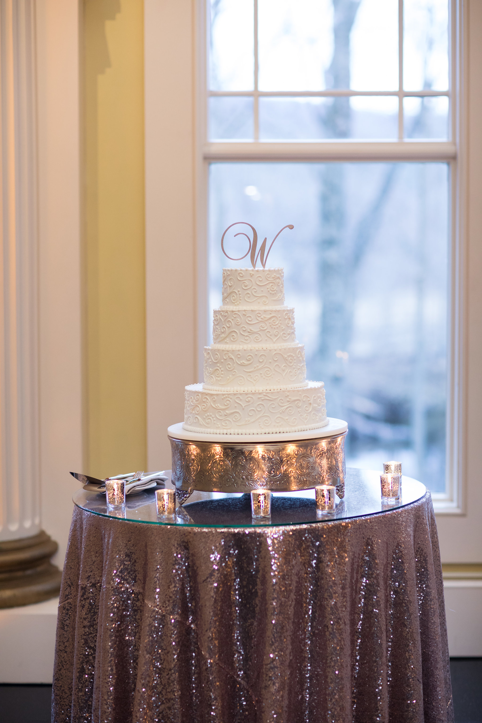 Four-tier white wedding cake with intricate piping and a "W" topper on a shimmering table.