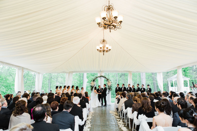 Wedding ceremony taking place under a draped white tent with chandeliers, featuring guests seated in rows facing the bride, groom, and wedding party at the altar.