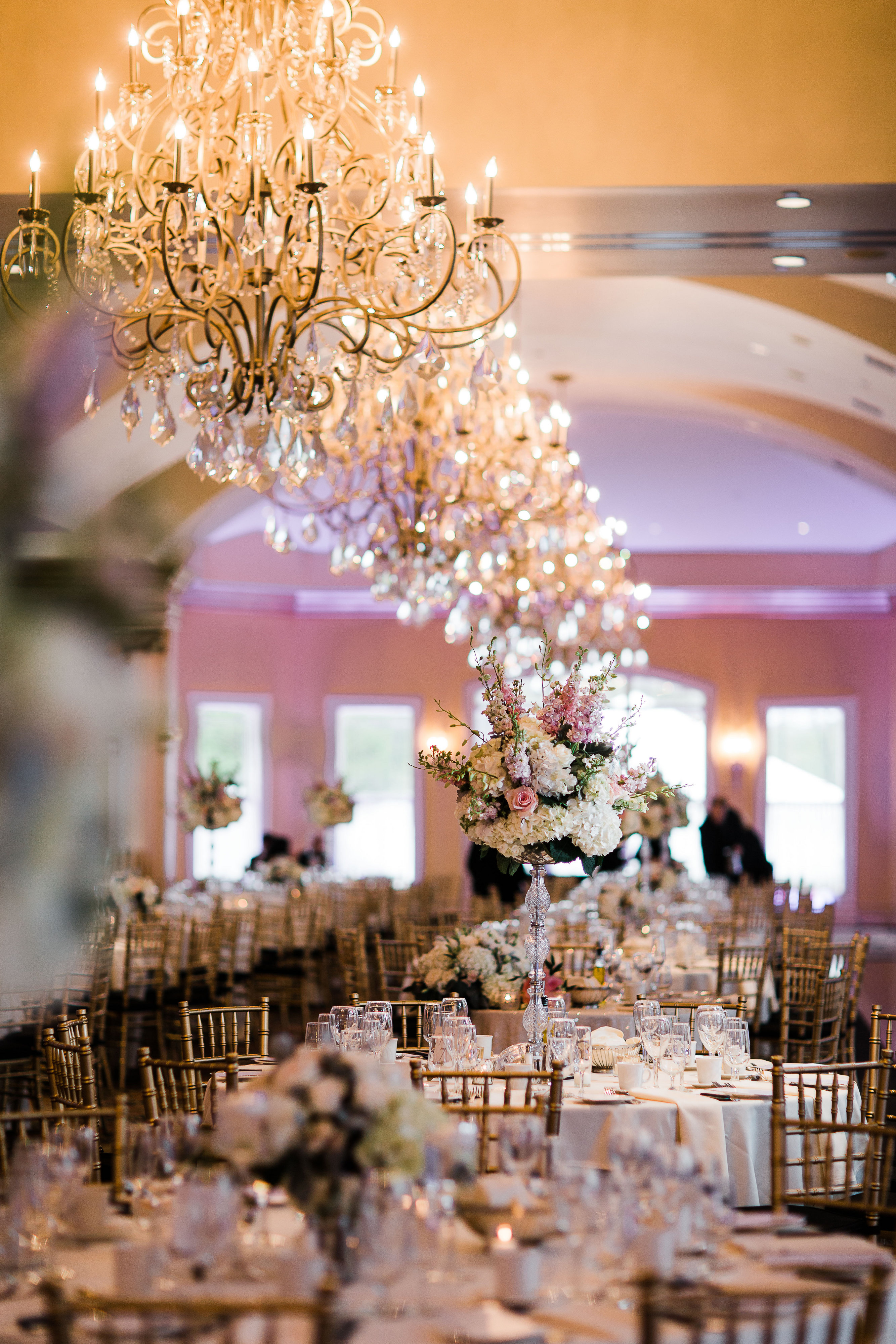 Elegant ballroom wedding reception with rows of crystal chandeliers hanging over tables set with floral centerpieces and gold chiavari chairs.