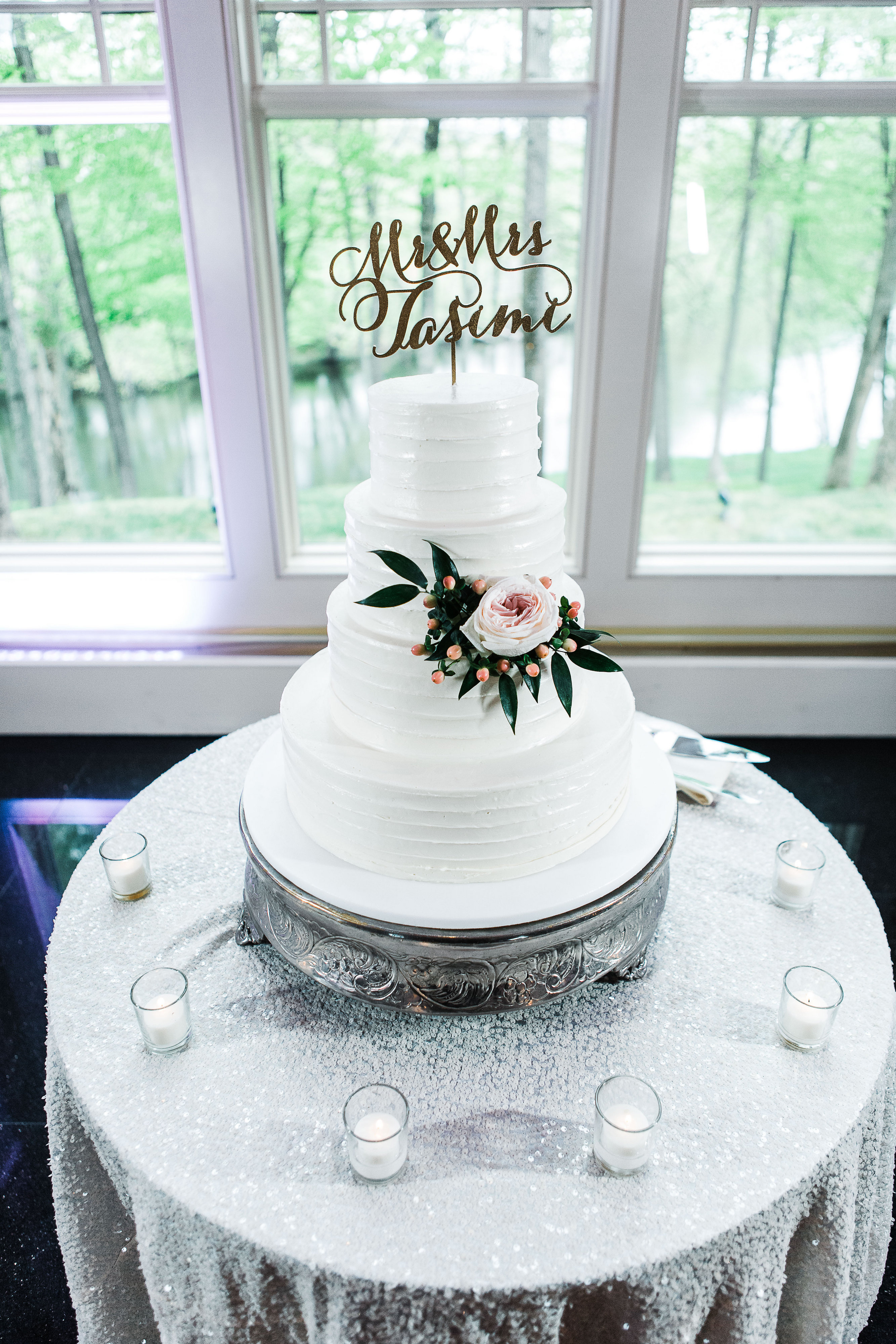 Four-tier white wedding cake with a "Mr. & Mrs. Tasimi" topper, decorated with a fresh rose.