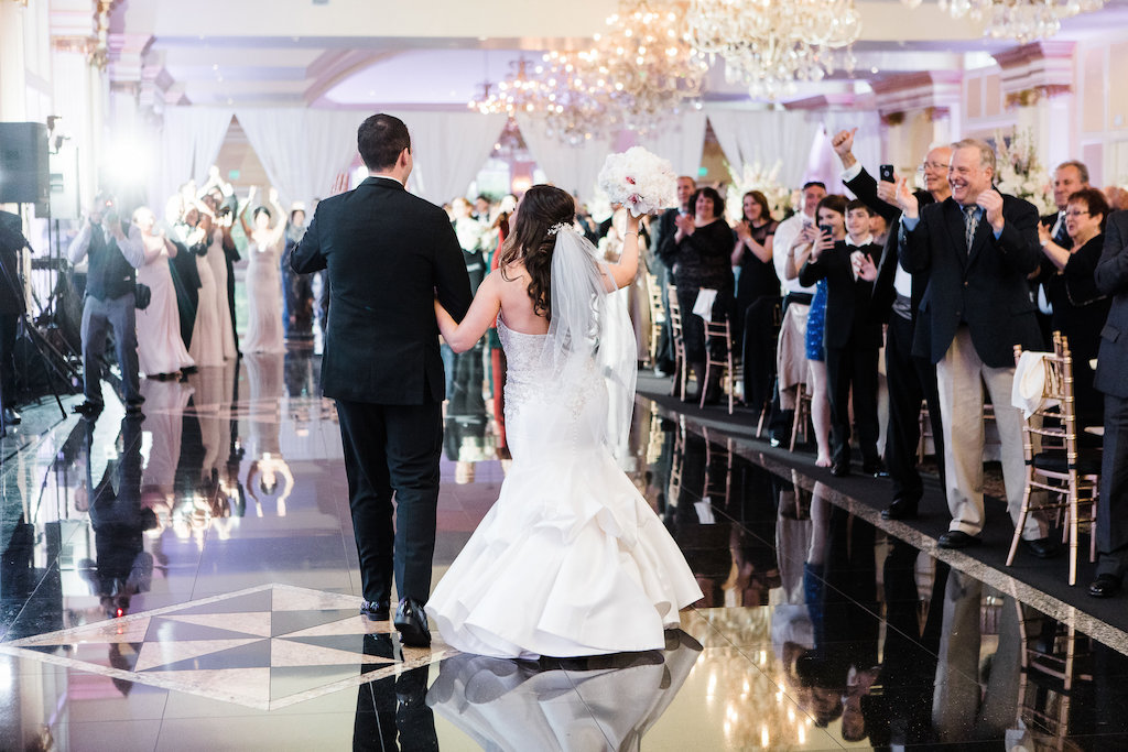 Bride and groom viewed from behind as they make their grand entrance into a wedding reception ballroom, cheered on by applauding guests.
