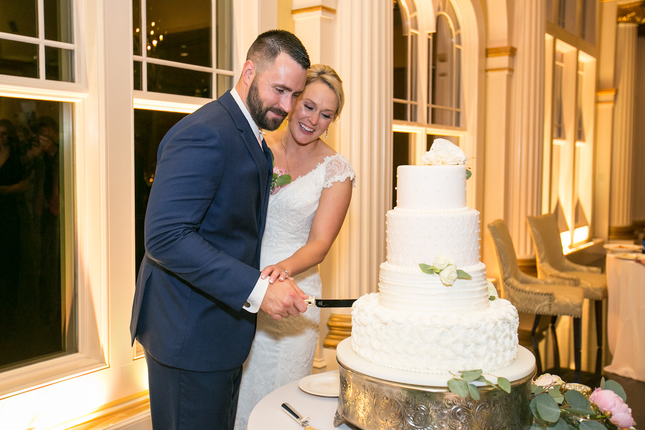 Bride and groom smile as they cut their four-tier white wedding cake together.