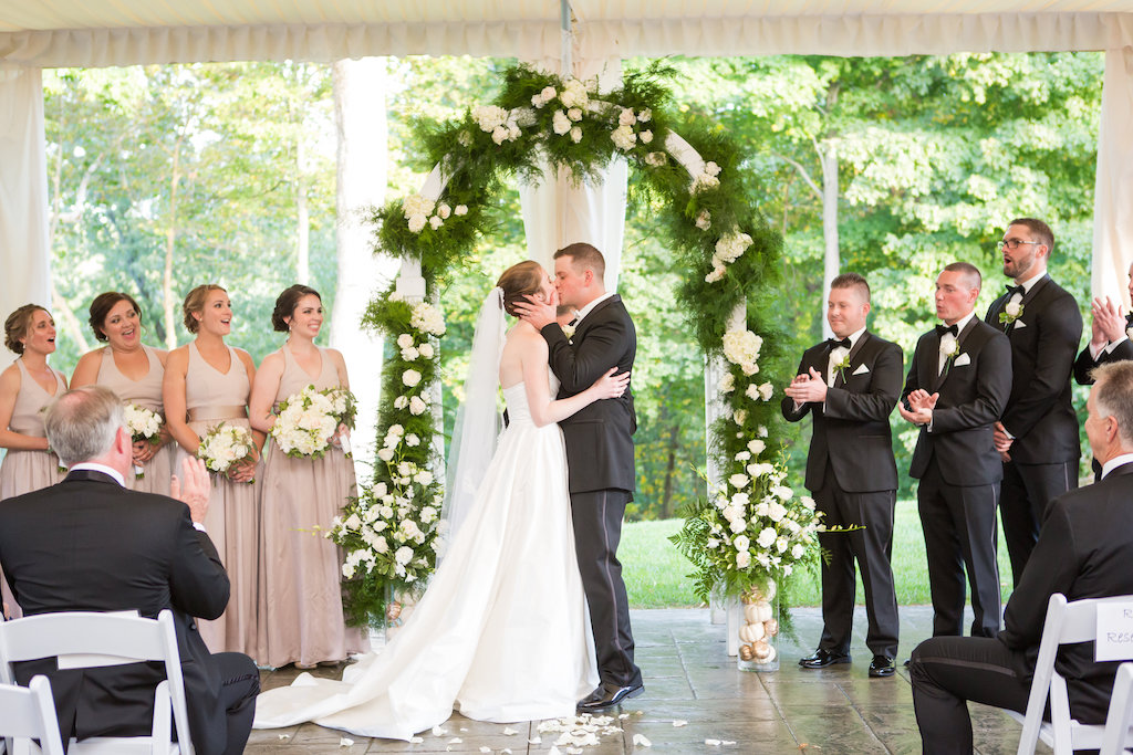 Bride and groom kissing under a greenery-covered wedding arch with their bridal party looking on.