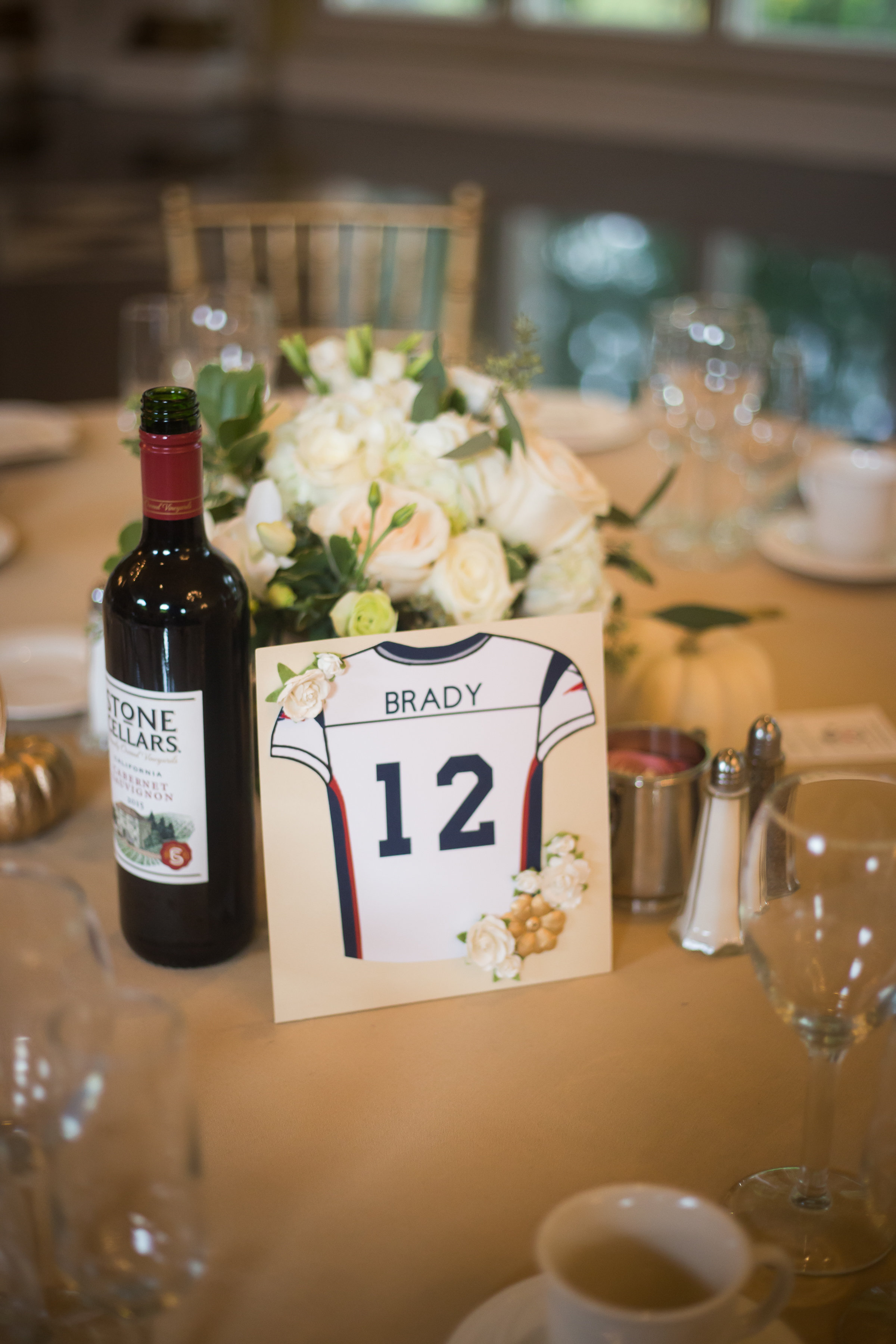 Wedding reception table setting featuring a wine bottle and a custom table sign shaped like a Tom Brady number 12 New England Patriots football jersey, decorated with small white floral accents.
