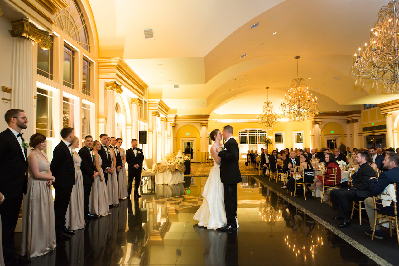 Bride and groom share their first dance on a reflective floor in a grand ballroom lined with guests.