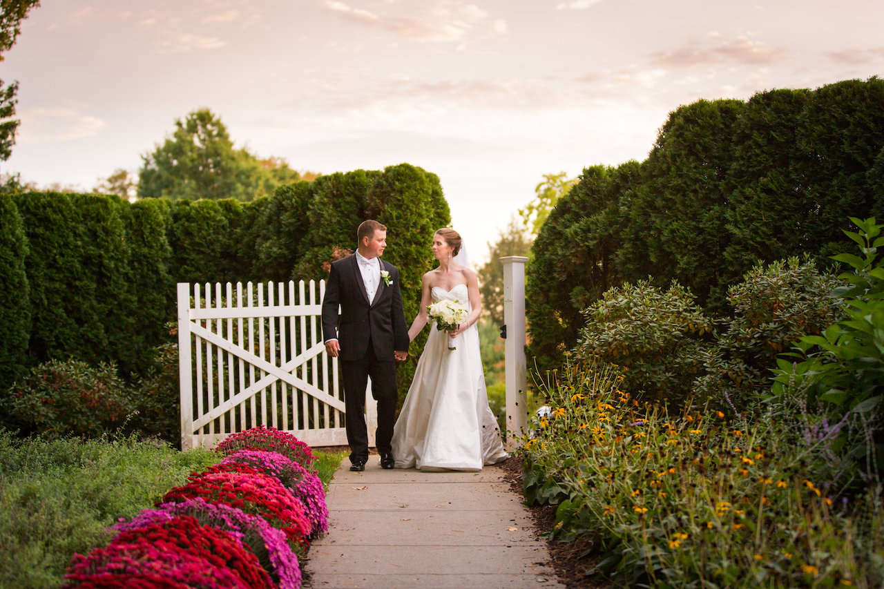 Bride and groom hold hands and walk down a garden path next to a white picket gate.