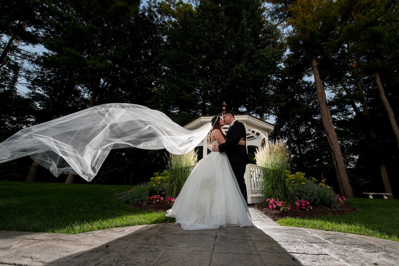 Bride and groom kissing in front of a white garden gazebo with the bride's long veil catching the wind.