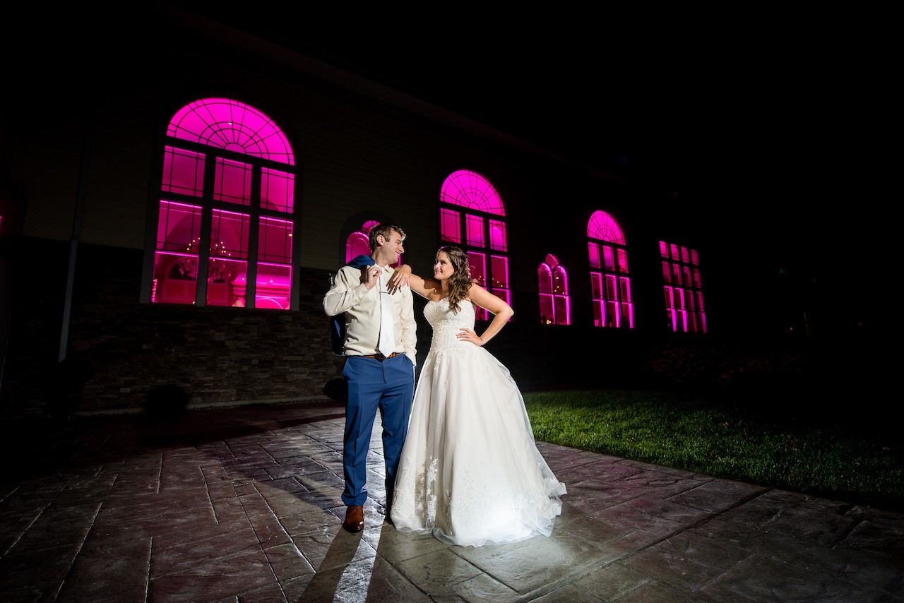 Newlyweds posing outside at night in front of a building with bright pink illuminated windows.