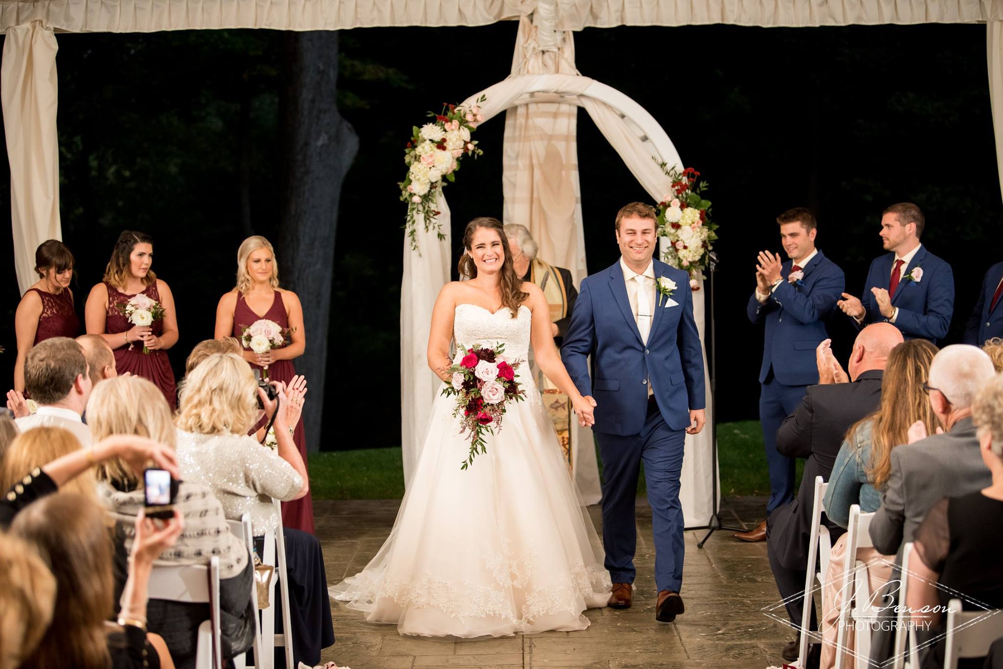 Newlyweds smiling and holding hands while walking down the aisle at an outdoor evening wedding ceremony under a decorated arch.