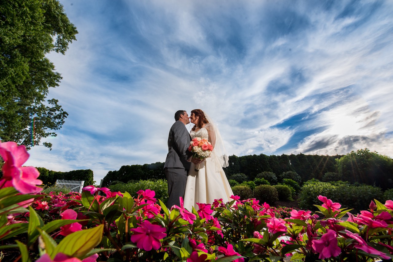 Bride and groom leaning in for a kiss while standing in a lush garden filled with vibrant pink flowers under a dramatic, cloudy blue sky.