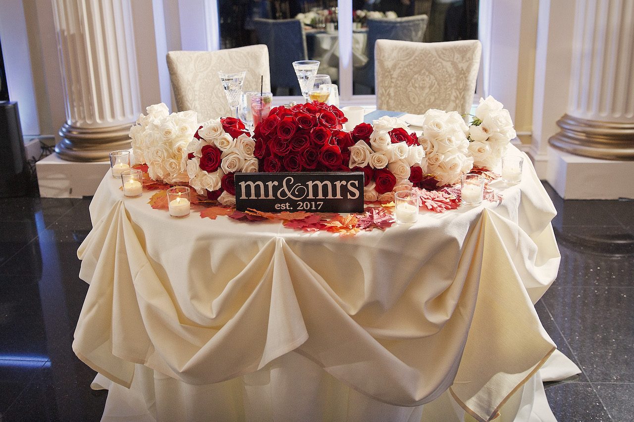 Sweetheart table for the bride and groom decorated with a "Mr. & Mrs. est. 2017" sign, bouquets of red and white roses, candles, and autumn leaves.