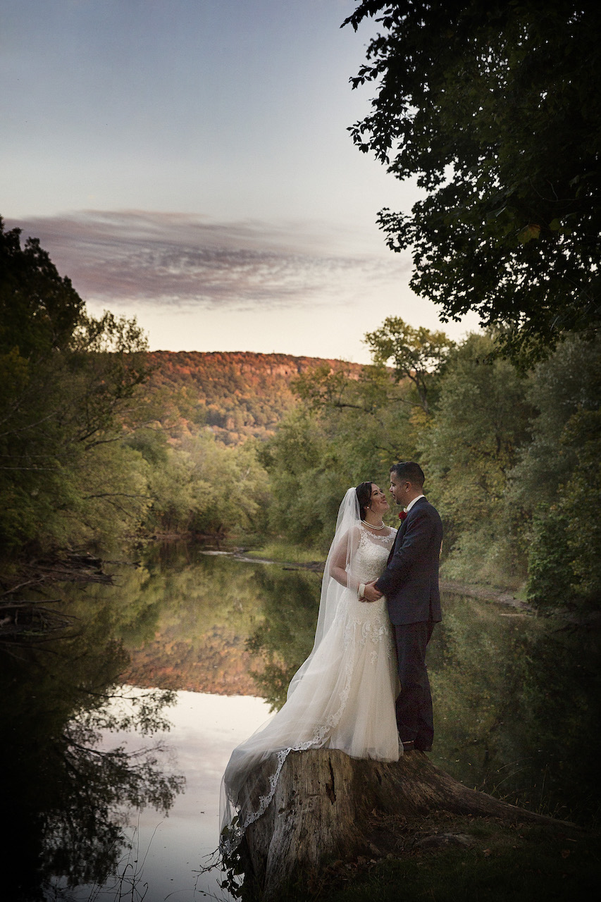 Bride and groom posing on a tree stump by a river reflecting autumn mountains at sunset.