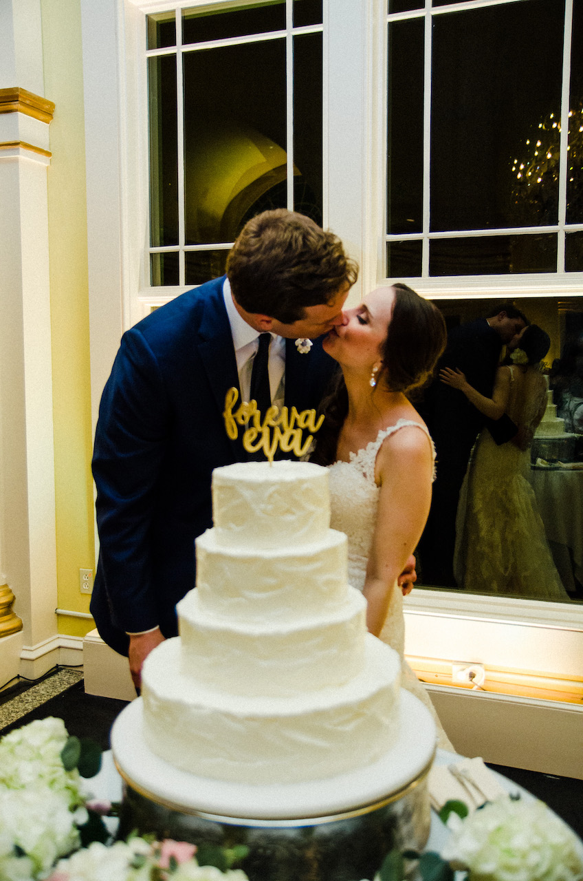 Bride and groom kiss behind a four-tier white wedding cake with a "foreva eva" script topper.