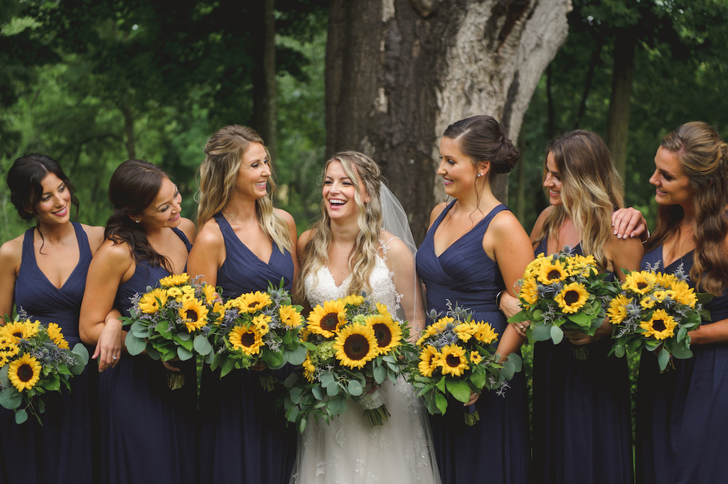 Bride laughing with her bridesmaids in navy blue dresses, all holding vibrant sunflower bouquets.