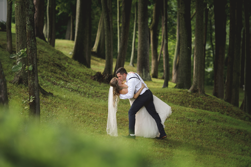 Groom dipping and kissing his bride on a grassy slope in a wooded forest setting.