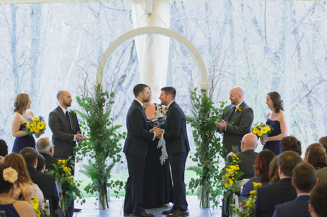 Two grooms holding hands at the altar during a wedding ceremony under an arch with greenery.