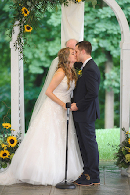 Bride and groom share their first kiss during an outdoor wedding ceremony under a white arch decorated with sunflowers.