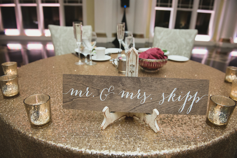 Wooden "Mr. & Mrs. Skipp" sign on a gold sequin-covered sweetheart table at a wedding reception.