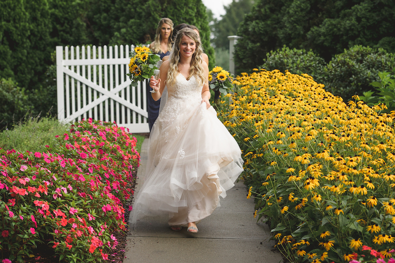 Bride walks down a garden path lined with yellow and pink flowers, followed by her bridesmaids.
