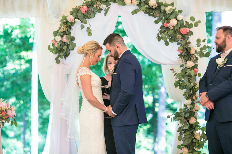 Bride and groom hold hands with eyes closed during a wedding ceremony under a floral arch.