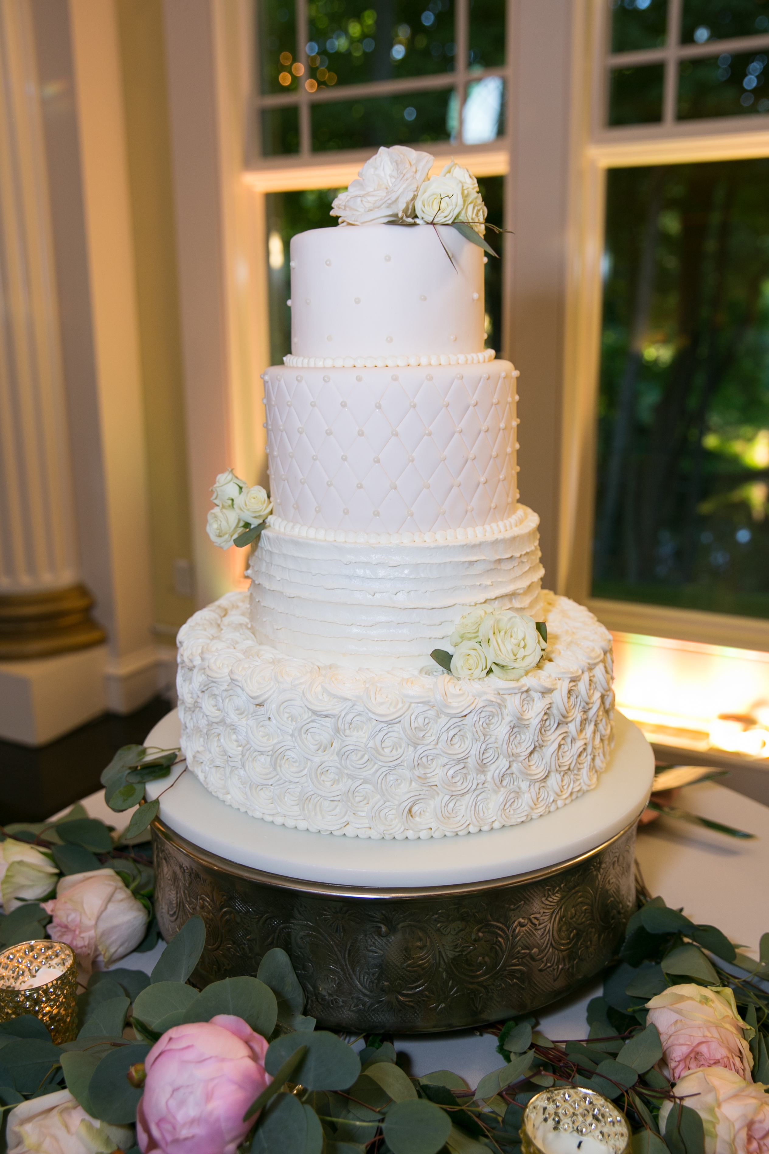 Four-tier white wedding cake featuring a different texture on each layer, including swiss dot piping, a quilted pattern, horizontal ruffles, and a bottom rosette layer, decorated with white roses and eucalyptus leaves.