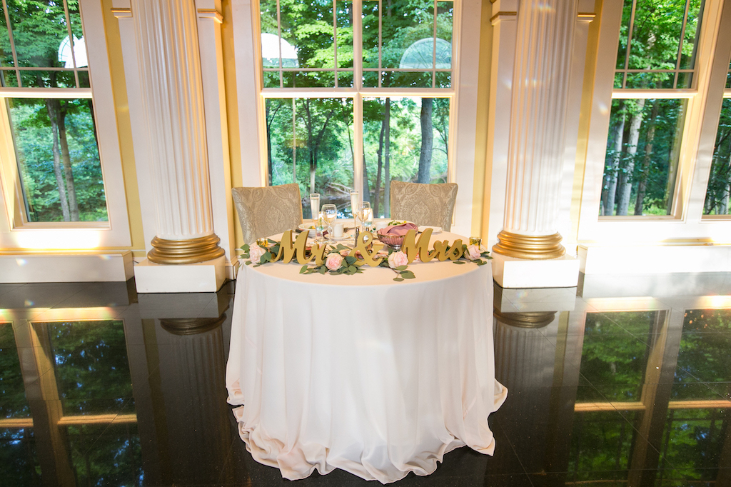 Sweetheart table for the bride and groom decorated with a "Mr. & Mrs." sign and pink roses, positioned in front of a scenic window.