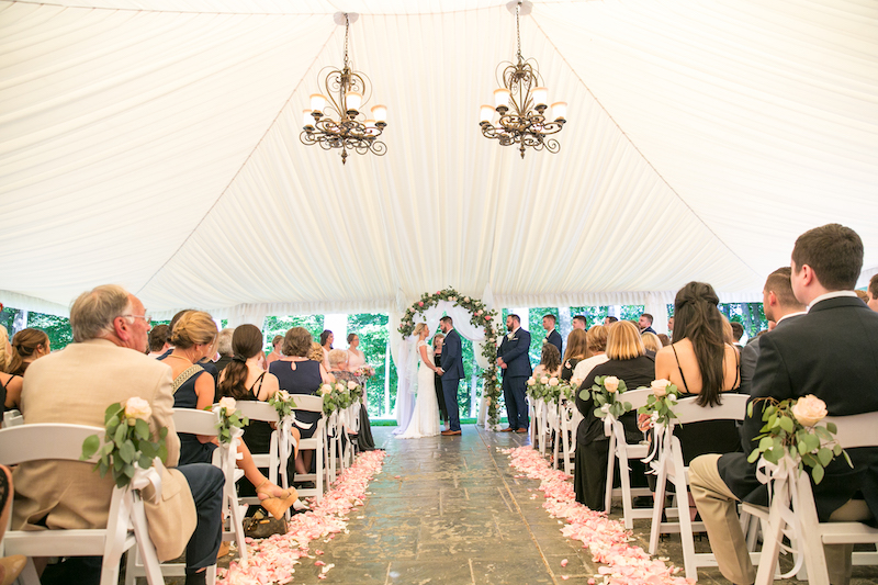 Bride and groom at the altar during a tented wedding ceremony with guests seated on both sides.