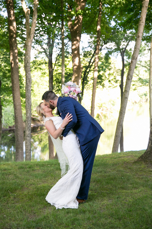 Groom dips and kisses his bride on a grassy lawn in front of trees and a river.