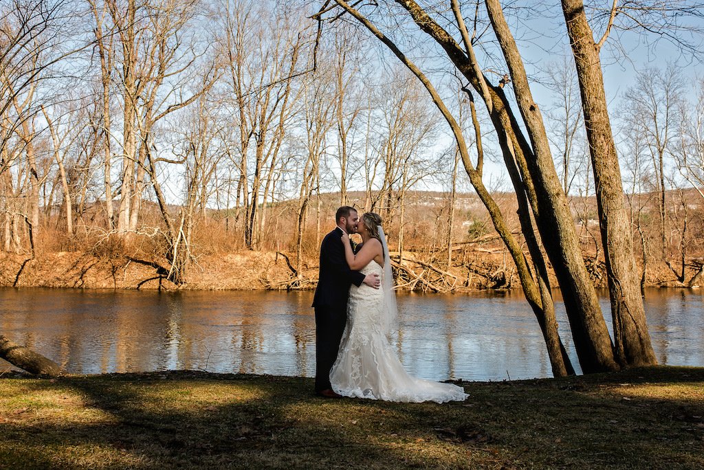 Bride and groom kissing by a scenic riverbank lined with bare autumn trees.