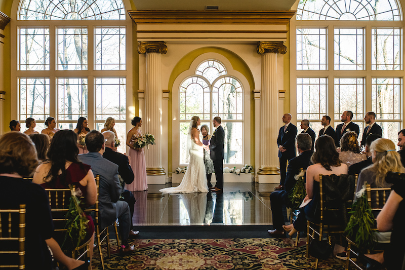 Bride and groom stand at the altar during an indoor wedding ceremony with tall arched windows.