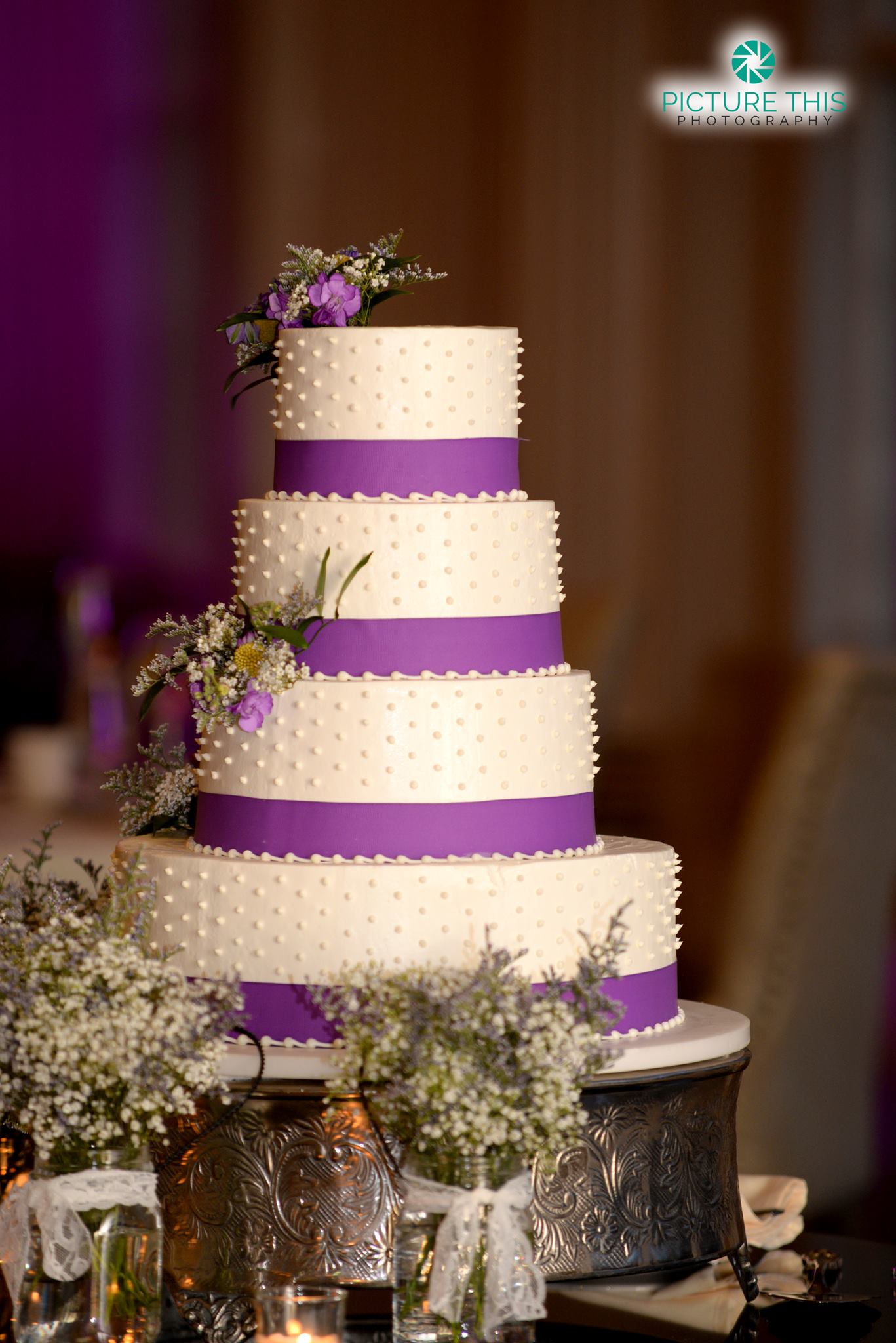 Four-tier white wedding cake with swiss dot piping and purple ribbon bands, decorated with fresh purple and white flowers.